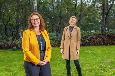 Twee dames staand op een grasveld kijken lachend in de lens 