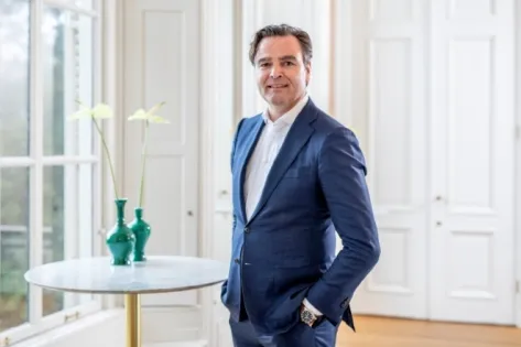 Zakelijk portret van een man in blauw pak die in een lichte kantoorruimte staat naast een tafel met twee groene vazen, passend bij een interview over de groei en successen van Gom in 2018.