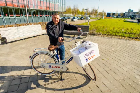 Medewerker met Gom-fiets op Schiphol, voorzien van mand met schoonmaakmaterialen, gebruikt voor efficiënte schoonmaak van bedrijfsgebouwen op de luchthaven.