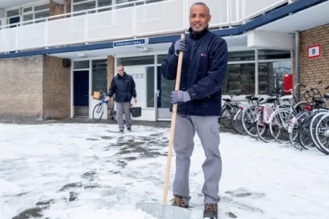 Twee medewerkers van Gom staan buiten bij een appartementencomplex op een besneeuwd trottoir. De voorste medewerker glimlacht en houdt een sneeuwschep vast, terwijl een collega op de achtergrond met een schop werkt. Op de achtergrond staan geparkeerde fietsen en een bord met de letter ‘B’. De foto benadrukt hoe Gom-medewerkers hun werk voortzetten onder winterse omstandigheden.