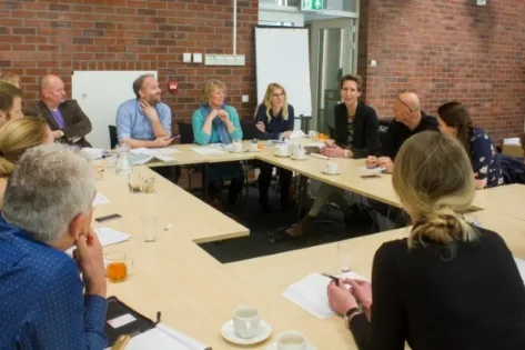 een grote groep van ruim 13 personen zit rondom een vergadertafel