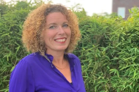 Portret van een vrouw met krullend haar en een paarse blouse, lachend voor een groene haag.