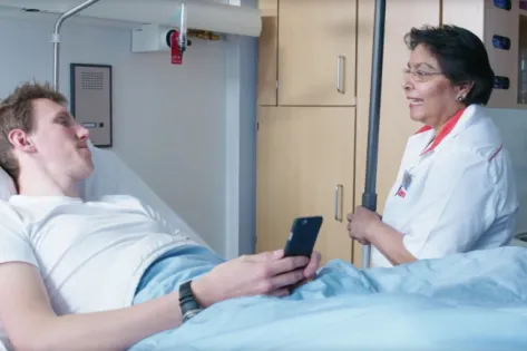 Schoonmaakmedewerker van Gom Zorg in gesprek met een patiënt in een ziekenhuisbed tijdens het schoonmaken van de kamer in Dijklander Ziekenhuis Hoorn.