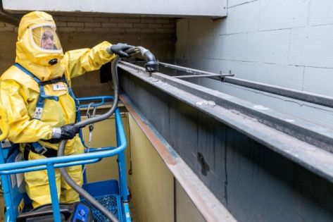 een medewerker van Gom Specialistische reiniging in een geel en geheel afgesloten beschermend pak zuigt gevaarlijke stoffen op.