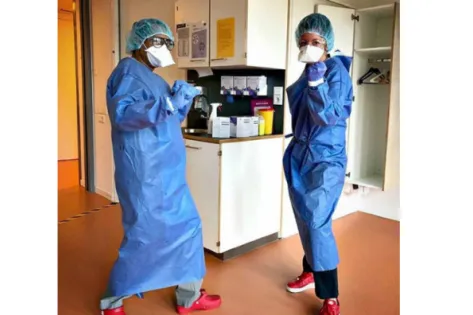 Twee zorgmedewerkers in blauwe beschermende kleding, mondmasker, handschoenen en haarnetje poseren in een ziekenhuiskamer.