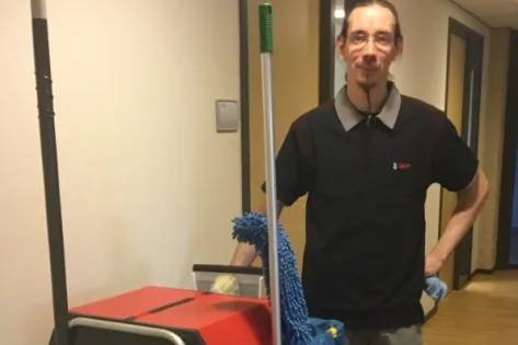 Schoonmaker in Gom-uniform poseert met schoonmaakkar en dweil in een gang van een kantoorgebouw.