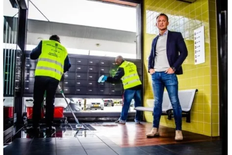 Twee schoonmakers in gele veiligheidshesjes maken een ruimte schoon, terwijl een man in pak en spijkerbroek toekijkt bij een muur met gele tegels.