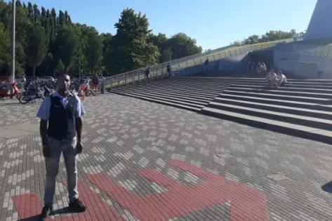 Beveiliger staat buiten op een plein met trappen bij een modern gebouw op een zonnige dag.
