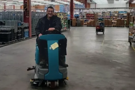 Man rijdt op een schrobmachine door een grote opslagruimte met rekken en stapels dozen tijdens schoonmaakwerkzaamheden.
