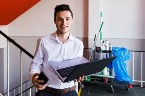 Mbo-student tijdens de facilitaire vakwedstrijd Skills Heroes met laptop en telefoon in een praktijkruimte met schoonmaakmaterialen op de achtergrond.