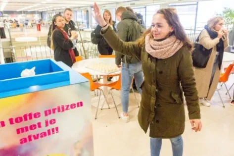 Student gooit afval in de kleurrijke interactieve prullenbak met de tekst ‘Win toffe prijzen met je afval!’ in een schoolkantine, terwijl andere studenten toekijken en meedoen.