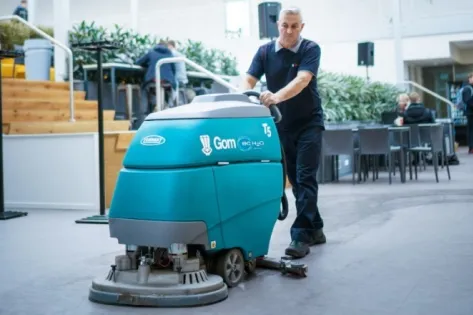 Medewerker van Gom bedient een schrobmachine tijdens het schoonmaken van de vloer in een grote binnenruimte.