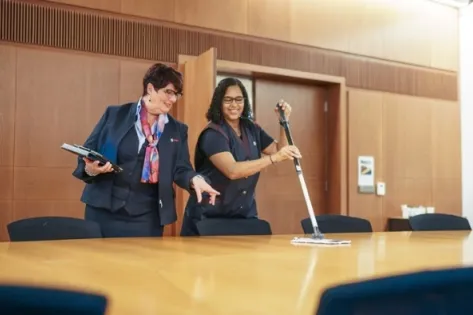 Twee Gom-medewerkers in uniforme bedrijfskleding werken samen in een vergaderruimte, waarbij één medewerker de tafel poetst en de ander aanwijzingen geeft met een tablet, als illustratie van opleiding en professionalisering in de schoonmaakbranche.