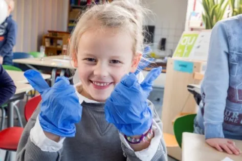 Kind in klaslokaal lacht en steekt duimen op terwijl het blauwe schoonmaakhandschoenen draagt, onderdeel van de proef met probiotica voor schonere schooltoiletten.