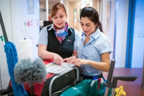 Twee Gom-medewerkers in uniform bespreken schoonmaaktaken aan een schoonmaakkar in een gang, terwijl ze samen een checklist doornemen.