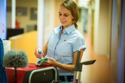 Een medewerker van Gom in zorgkleding maakt schoon in een ziekenhuisgang. Ze glimlacht terwijl ze schoonmaakmiddelen op een doek aanbrengt op haar werkkar, waarop diverse schoonmaakbenodigdheden staan zoals borstels en sprays.