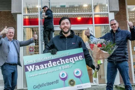 Blije medewerker ontvangt bloemen en een grote waardecheque van €500 tijdens de Gom Uitblinker Verkiezing, omringd door collega’s die applaudisseren en duimen opsteken.
