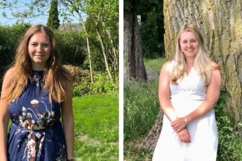 Twee jonge vrouwen staan buiten in een groene omgeving, de een in een donkerblauw bloemenjurk en de ander in een witte jurk.