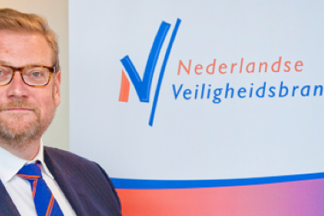 Man in pak staat voor een muur met daarop het logo van de Nederlandse veiligheidsbranche