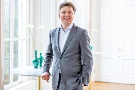 Man in grijs pak staat glimlachend in een lichte kamer met witte muren en groene vazen op de achtergrond.