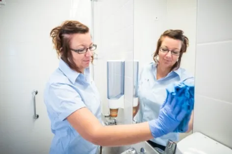 Schoonmaakmedewerker in lichtblauw uniform en handschoenen maakt een spiegel schoon in een sanitaire ruimte.