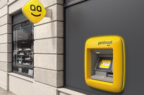Een gele geldautomaat hangt aan een grijze muur naast een winkel.