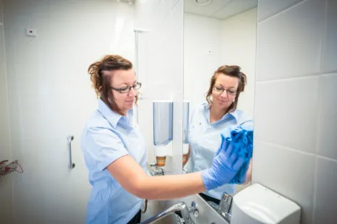 Schoonmaakmedewerkster van Gom poetst een wastafel in een zorginstelling en kijkt in de spiegel, als onderdeel van het gebruik van probiotica voor een frisse en hygiënische omgeving.