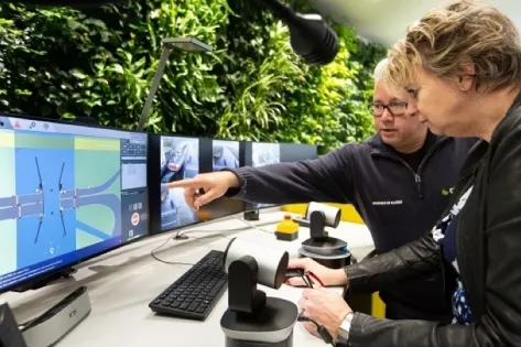 Twee medewerkers bekijken verkeersbeelden op meerdere monitoren in een controlekamer. Eén persoon wijst iets aan op het scherm terwijl de ander meeluistert.