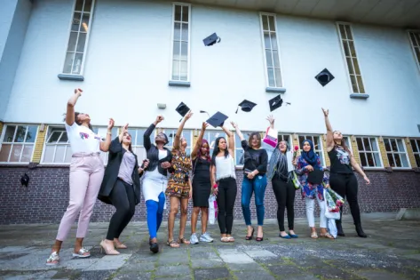 Een groep jonge vrouwen viert hun afstuderen buiten voor een schoolgebouw. Ze lachen en gooien gezamenlijk hun afstudeerhoedjes de lucht in, terwijl enkelen bloemen en diploma’s vasthouden. De sfeer is vrolijk en feestelijk.