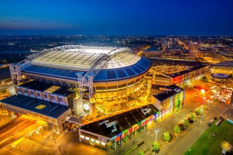De kijk van bovenaf op de  Johan Cruijff Arena in Amsterdam in de avond waarbij zowel de arena als het treinstation is verlicht.