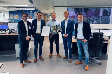 Vijf mannen in pak staan op de AlarmServiceCentrale geposeerd met een certificaat en bloemen in hun hand.