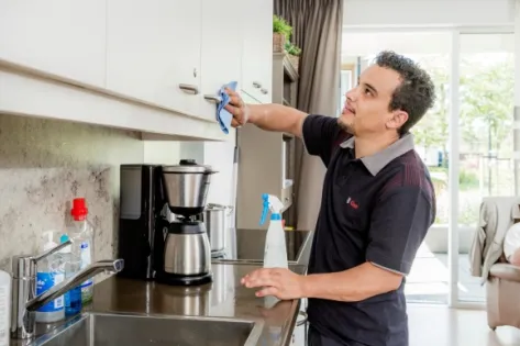 Schoonmaakmedewerker van Gom poetst keukenkastjes in een zorgwoning, terwijl een bewoner op de achtergrond in de woonkamer zit, als illustratie van ondersteuning in de zorgsector.
